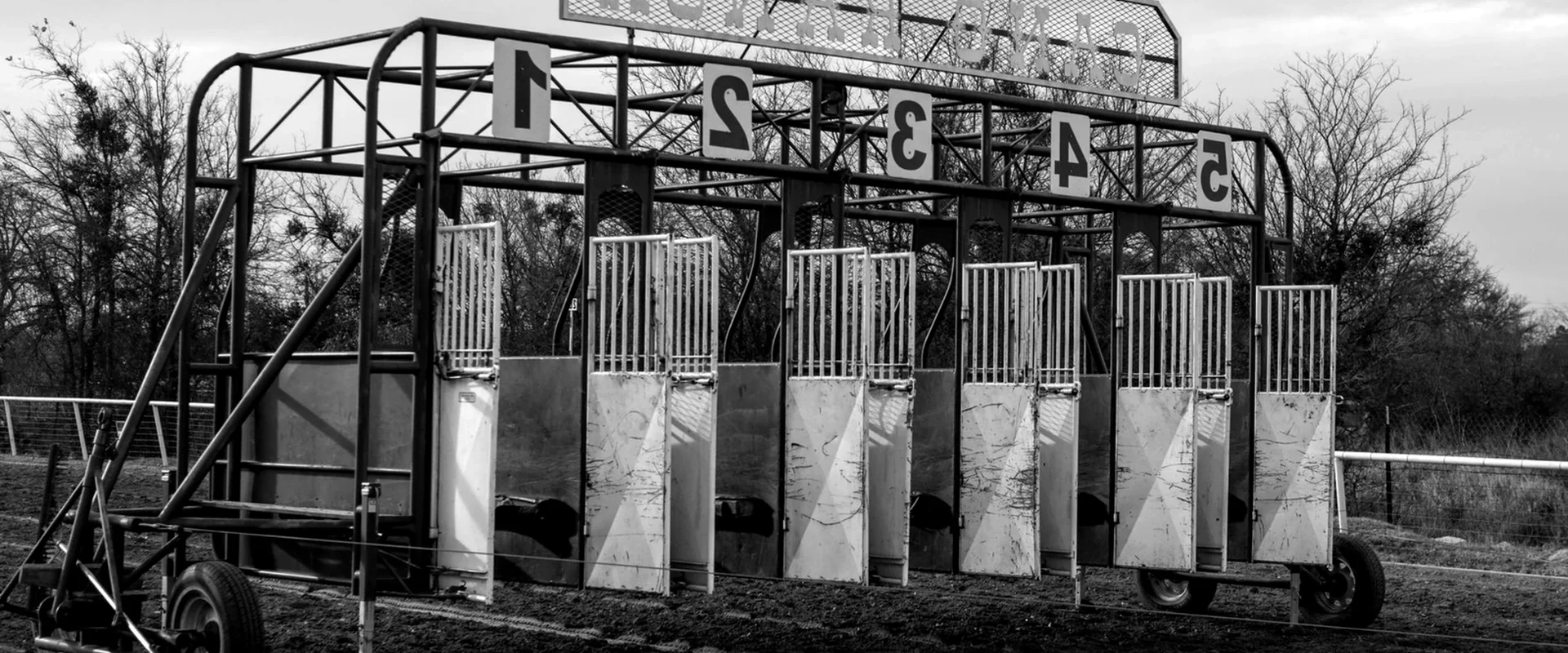 Black-and-white photograph of an empty mobile starting barrier on bare ground, its numbered bays standing open