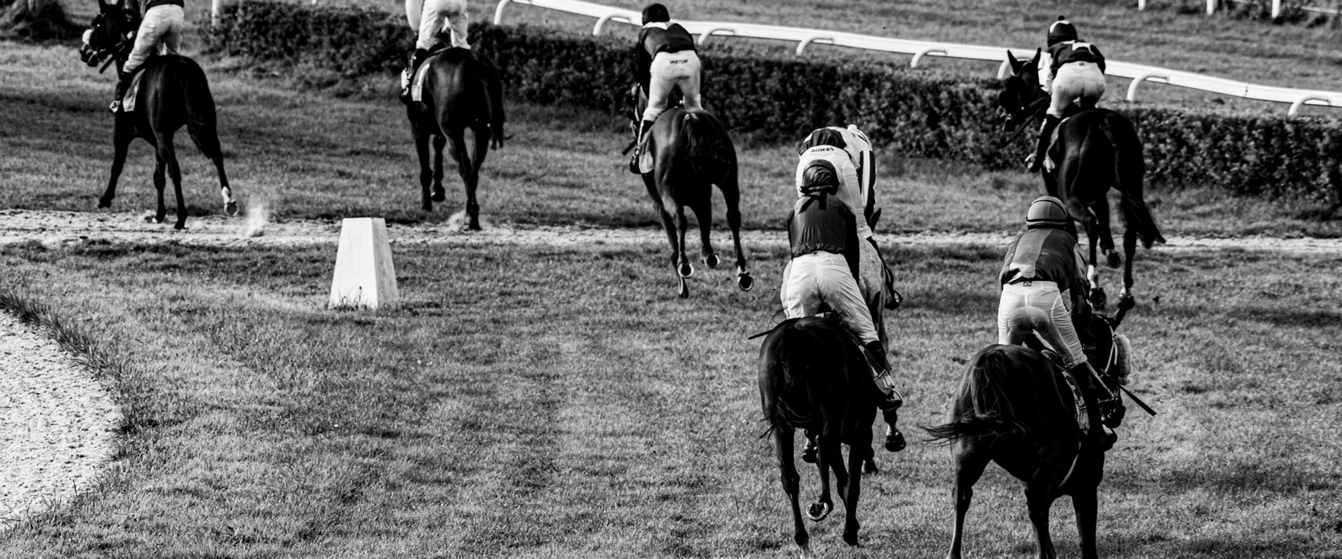 Black-and-white photograph of a thoroughbred field riding away from the camera on turf, the riders distinguishable only by their silks and caps